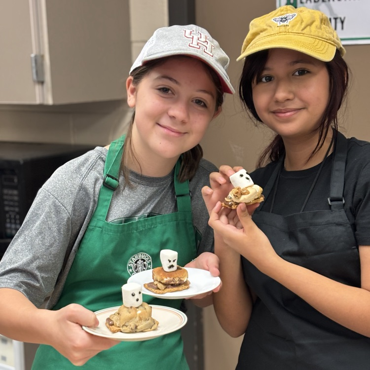 Two students wearing aprons, smiling. One holds two plates with cookies decorated with marshmallows.