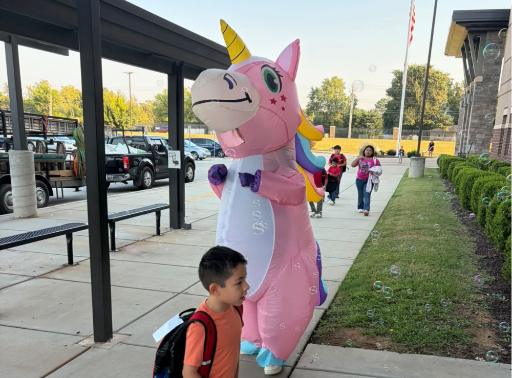 A teacher in an inflatable unicorn costume interacting with students during morning drop off