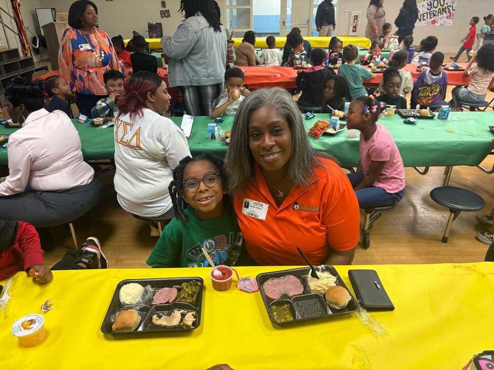 A parent and student sit together at a table with a lunch tray smiling, while other students and families share the cafeteria in the background.