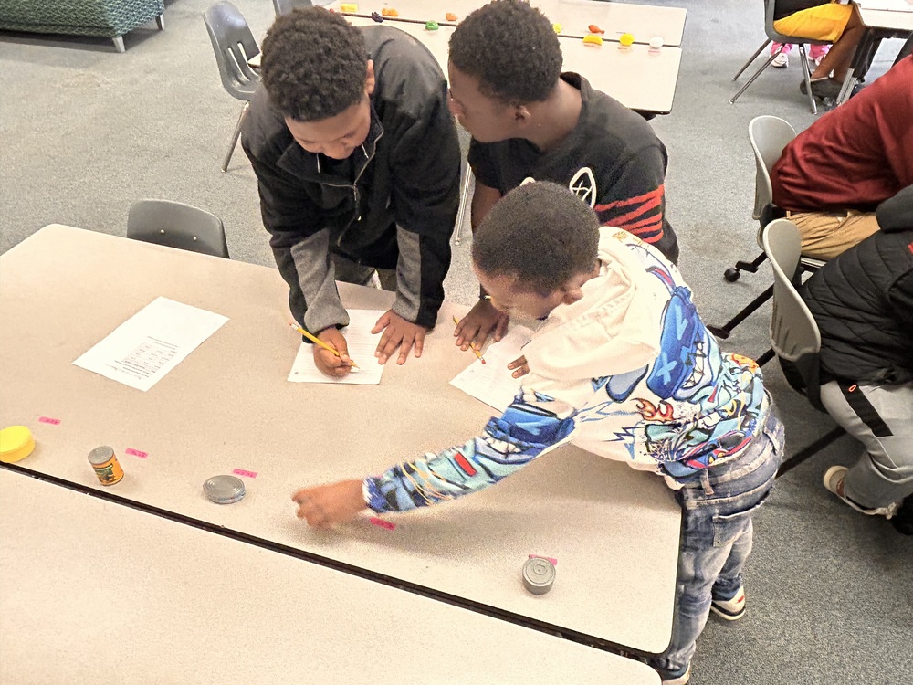 Students are gathered around play food options while calculating unit rates at a table in the library. 
