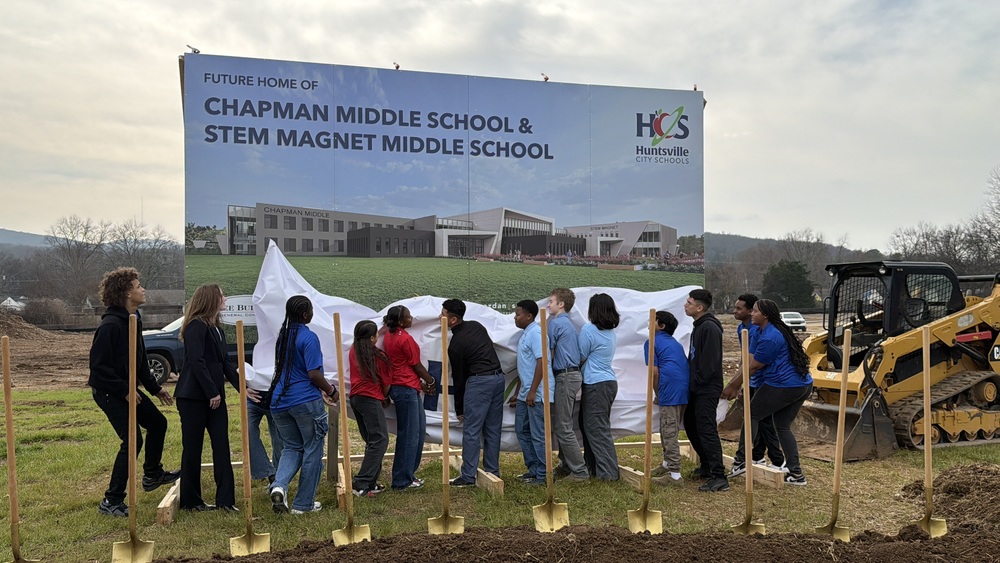 Student representatives pull back a white silk cover to unveil a large sign showcasing the architectural rendering of the future Chapman Middle and STEM Magnet Middle School campus. Gold shovels and construction equipment sit ready at the groundbreaking site.