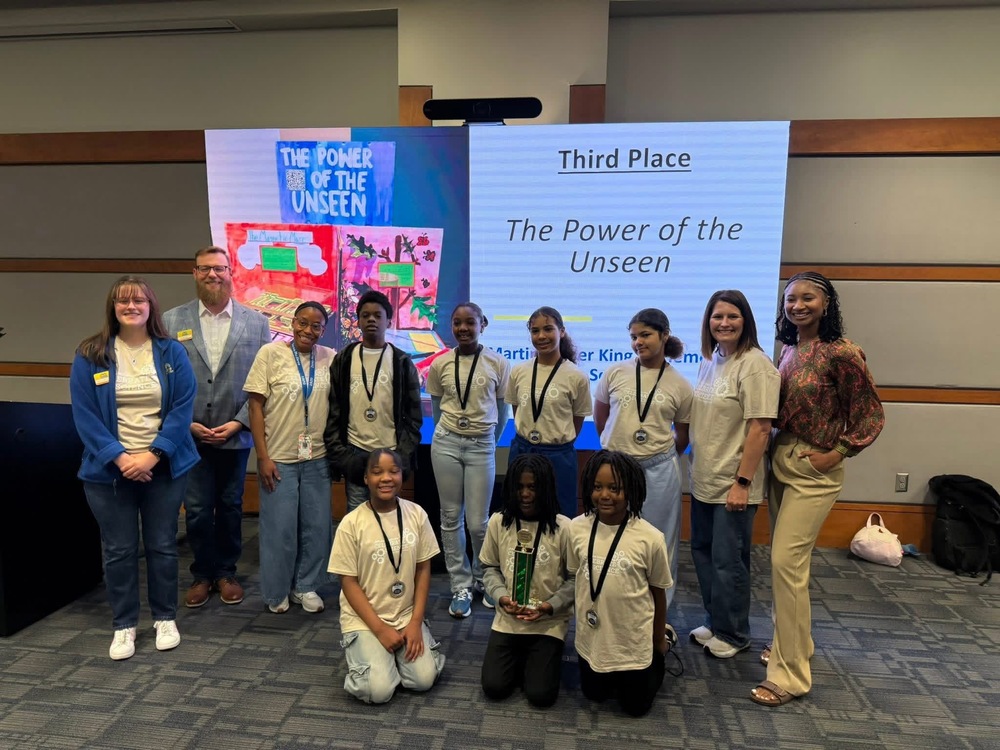 MLK students pictured in white shirt standing by volunteers at science competition. standing behind them is a screen that says the power of the unseen 