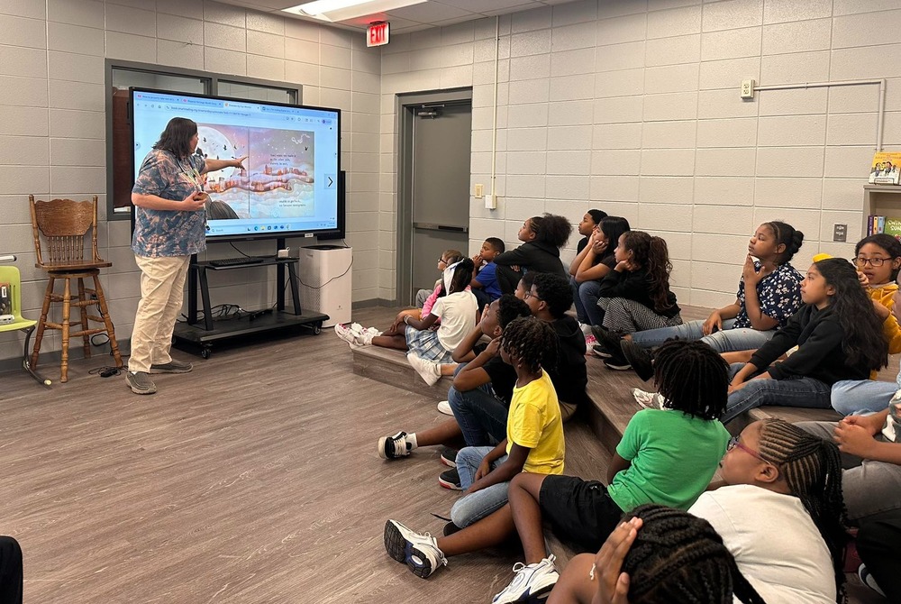 Mrs. Delacey stands at the smartboard displaying a story while students sit on the floor and listen