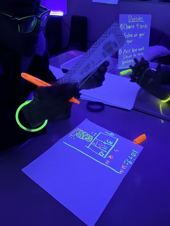 A student is sitting in a dark room, wearing glow bracelets, and solving math problems on white paper  using a highlighter.
