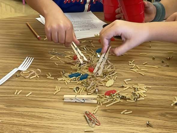Student hands are using a clothespin to pick up small noodles.