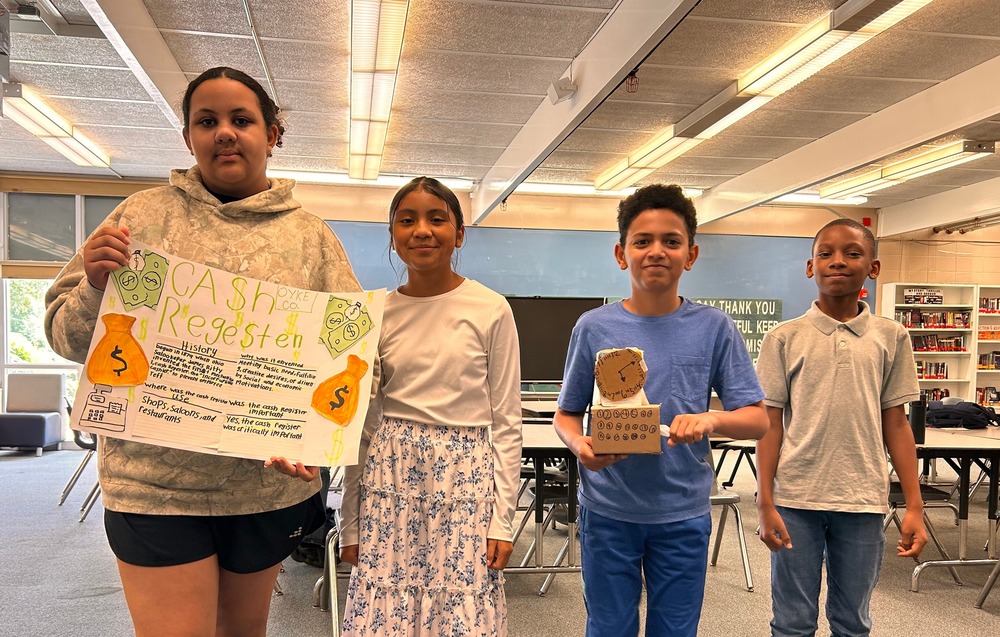 A group of students pose with their cash register prototype and informational poster for the Shark Tank Industrial Age competition in the library. 