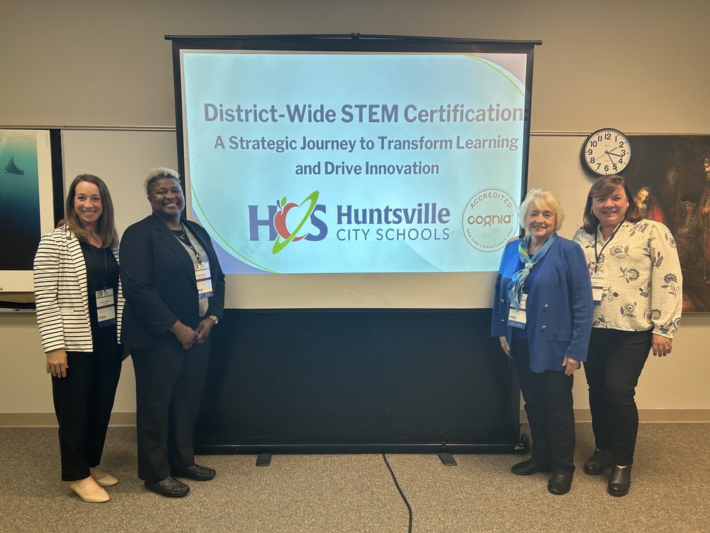 Rachel Evans, Bonnie Garrett, Cathy McNeal, and Rebecca Campbell at the Cognia Conference, Powerpoint slide behind them which reads District-Wide STEM Certification, a Strategic Journey to Transform Learning & Drive Innovation