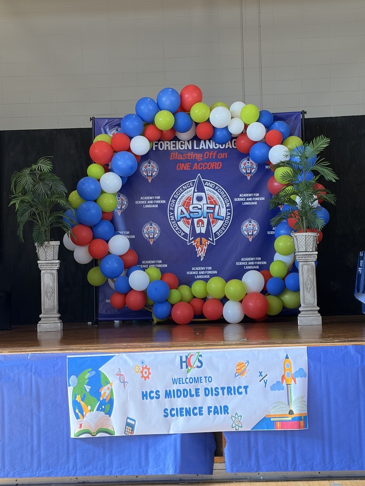 A stage decorated with a red, blue, green, and white balloon arch framing a "Foreign Language" banner, set above a "HCS Middle District Science Fair" welcome banner.