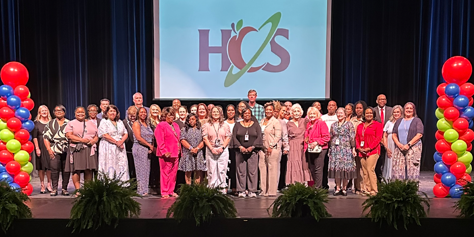Pinning Ceremony recipients stand on stage with Supt. Sutton and Deputy Supt. A.J. Buckner, with balloon columns and the HCS logo in the background