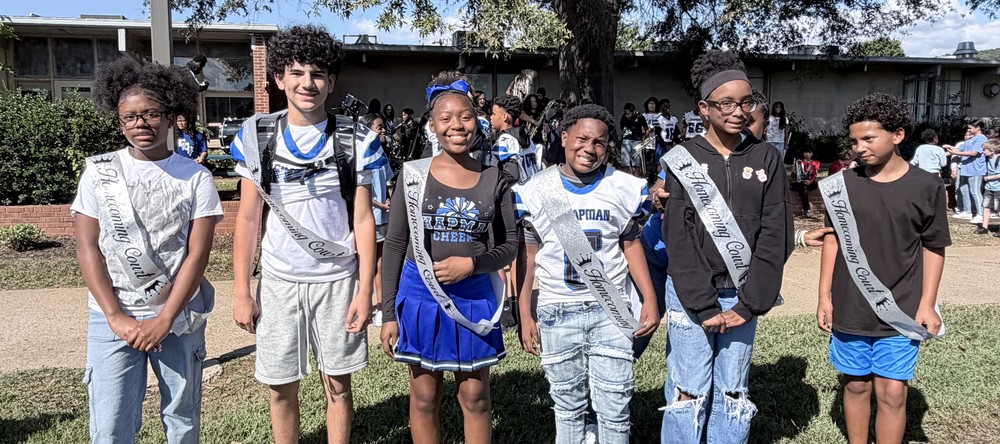 Chapman's 2025 Homecoming Court, a male and female representative from each grade level,  is lined up outside in the courtyard with the band playing in the background during the pep rally. 