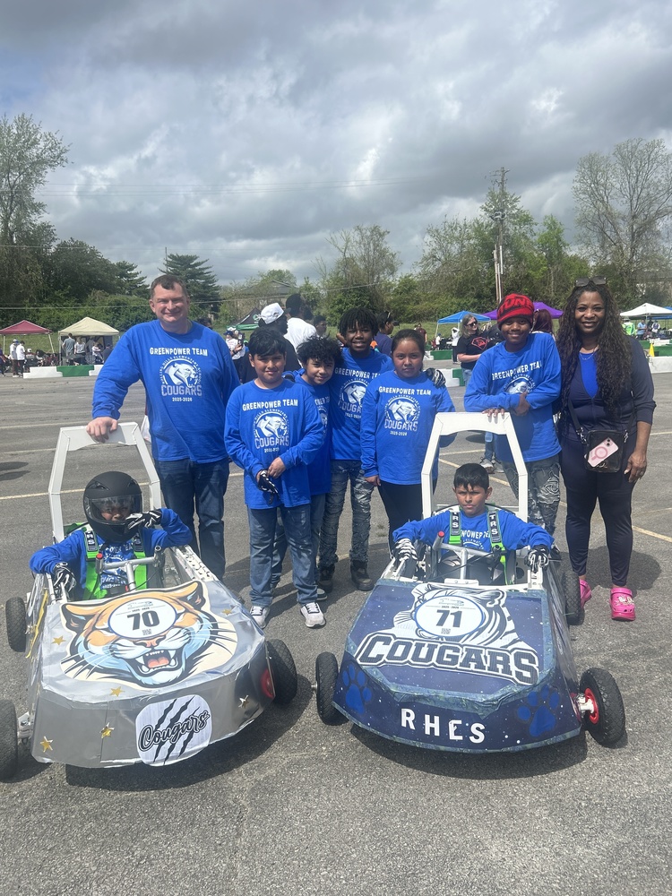 Students and teacher in matching blue shirts pose behind two cougar-themed soapbox cars at the HCS Greenpower Race.