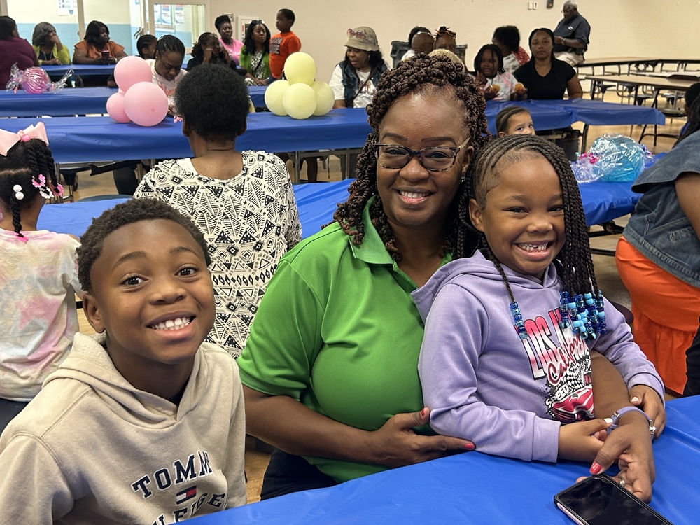 A smiling woman sits between two happy children at a decorated table with blue tablecloths and balloons, lin the cafeteria of the school . The room is filled with families and laughter, creating a warm and joyful atmosphere.