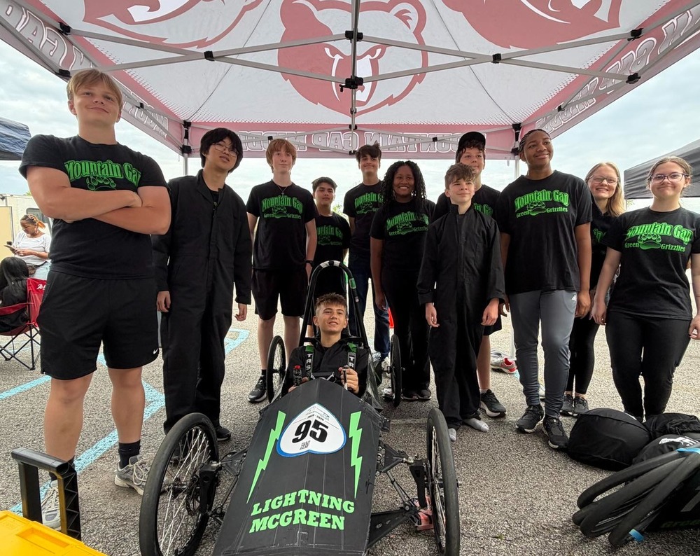 The mountain gap greenpower team standing behind their car and one student is sitting in the car. 