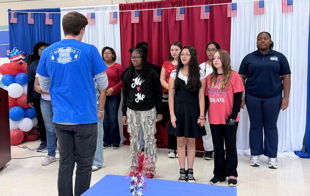 Members of Chapman Choir are standing in front of a red, white, and blue display for Veterans' Day as Mr. Hicks is leading the choir in song.