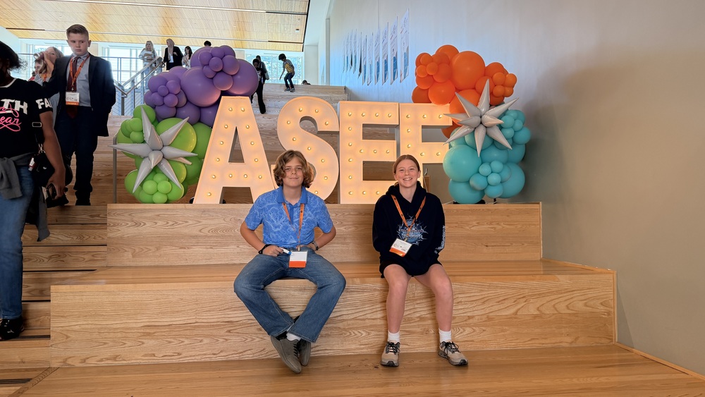 Hazel and Drew at the state science fair sitting on front of the Science Fair sign with balloons.
