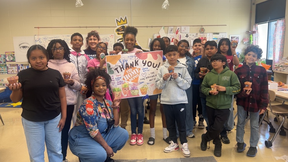 Art Club students pose together holding painted terra cotta pots and a thank-you sign in the classroom.