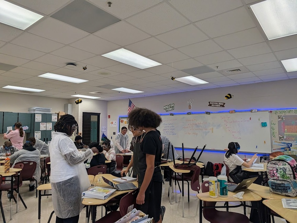 Students in science class standing and sitting around tables wearing jackets, safety glasses using tools collaborate on an assignment or experiment