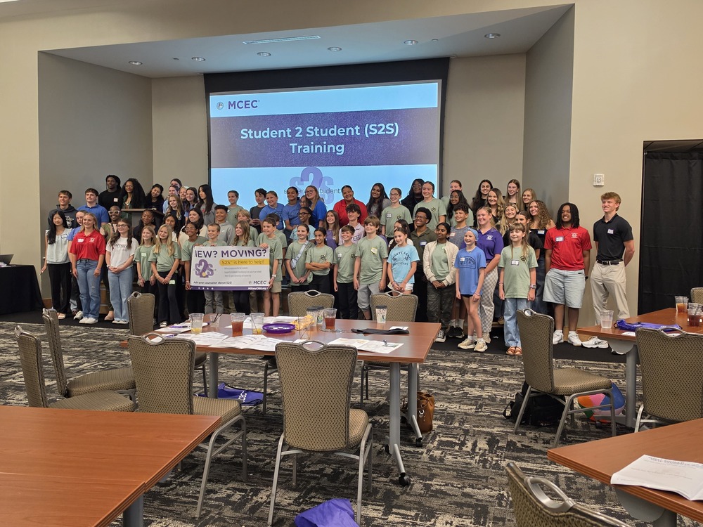 Columbia High School sudents from S2S at the summit with MCEC Student 2 Student sign up on white bord behind them while they stand holding up a banner posing for picture