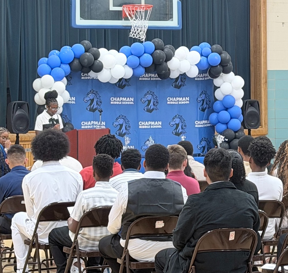 In the school gymnasium during a ceremony, where an audience sits in chairs facing a stage area. A speaker stands at a podium in front of a blue Chapman Middle School backdrop adorned with a large blue, black, and white balloon arch beneath a basketball hoop.
