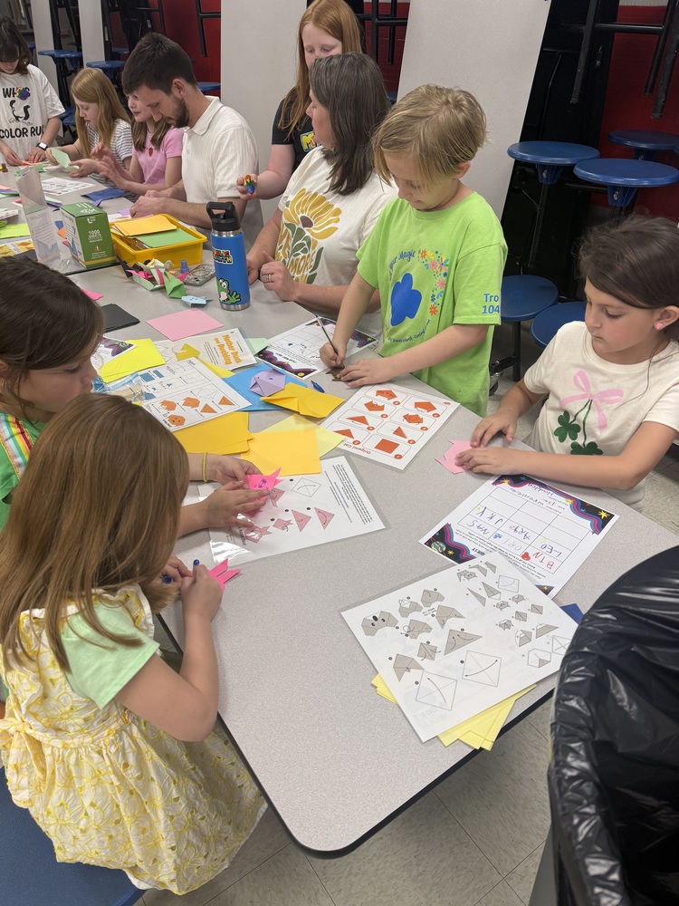 A group of children and adults working on origami at a table covered with colored paper and instructions.