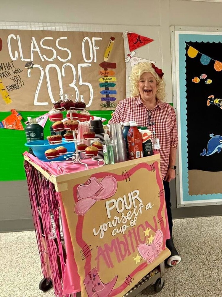 Principal Melillo poses behind a decorated “Pour yourself a cup of ambition” coffee cart in a classroom, surrounded by drinks and a “Class of 2035” display.