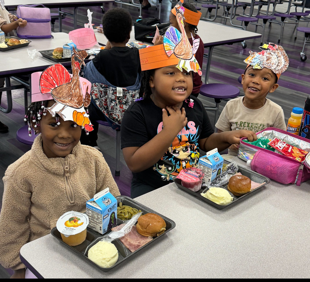 3 students are seated at a table, wearing  turkey headbands they colored, with their trays of food in front of them.