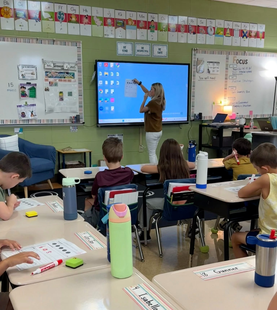 Ms. Moore in front of a first grade class giving instruction on the board.