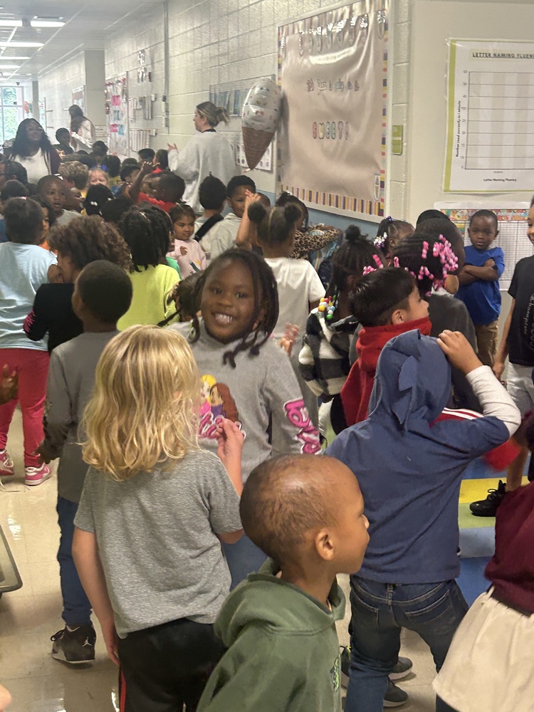 A group of young students are dancing excitedly in a brightly lit school hallway. Several children in the foreground are smiling with their arms raised, while others cheer and clap
