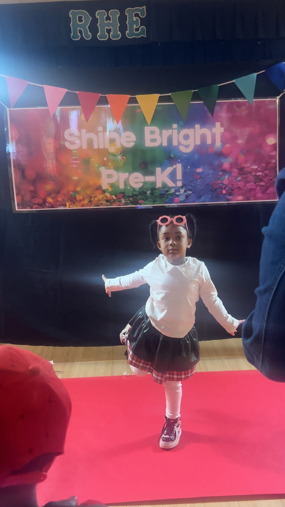 A Pre-K student poses on a red carpet in front of a colorful “Shine Bright Pre-K!” backdrop at RHE.S