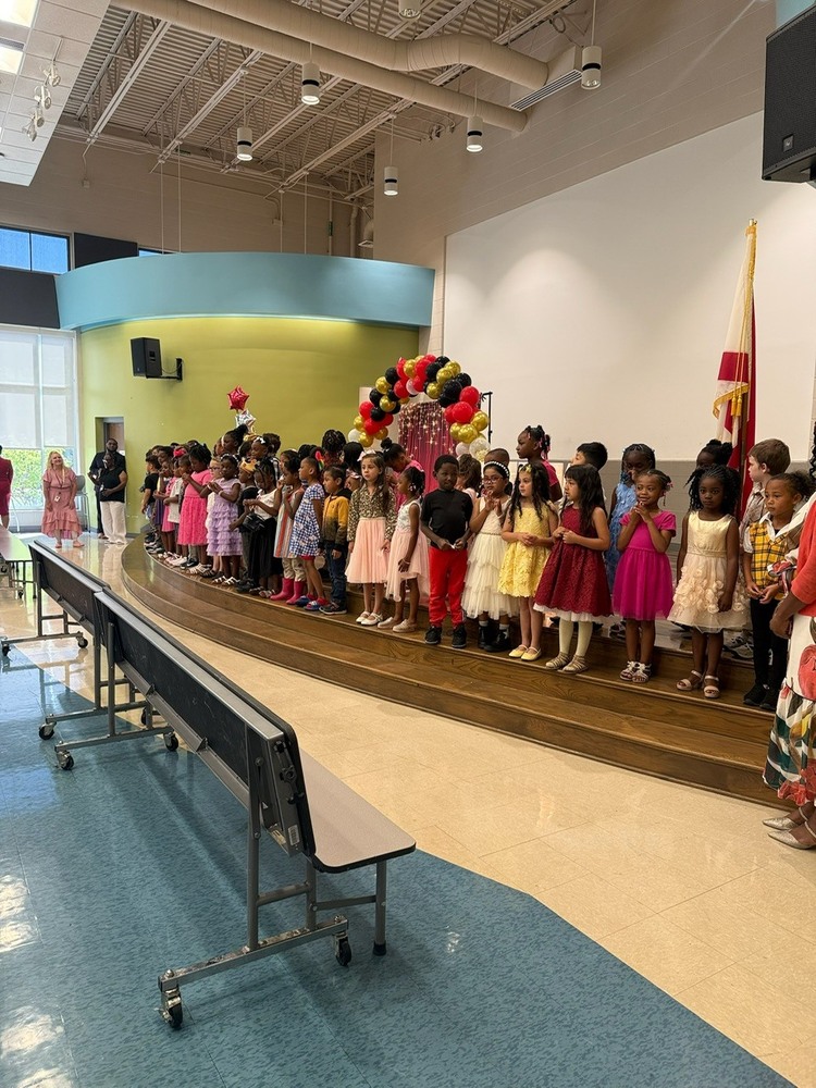 A large group of kindergarten students stands together on the stage during a Sonnie Hereford Elementary end-of-year celebration and awards ceremony. The students are dressed in colorful formal attire while families and staff watch proudly. Balloon decorations in red, black, and gold frame the stage, creating a festive atmosphere as the class celebrates their accomplishments and graduation milestone.