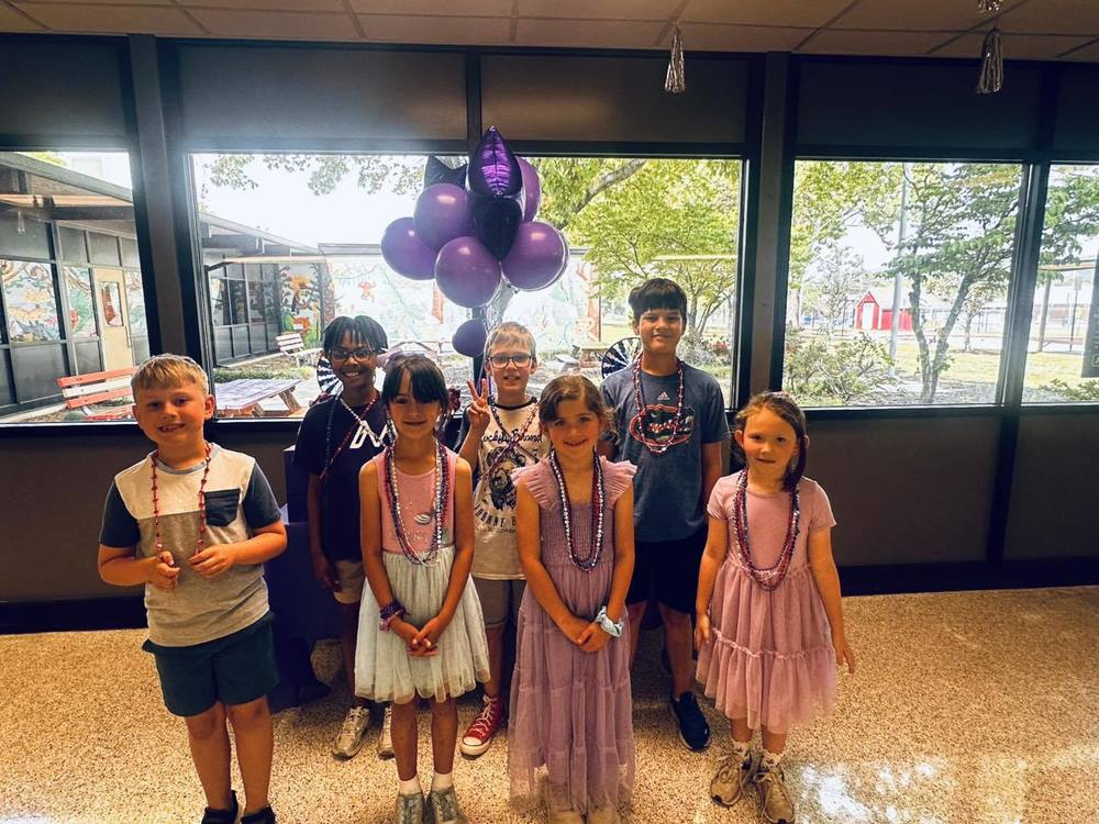 A group of seven smiling children wearing beaded necklaces stands in front of large windows with purple balloons behind them to celebrate Purple Up  Day.