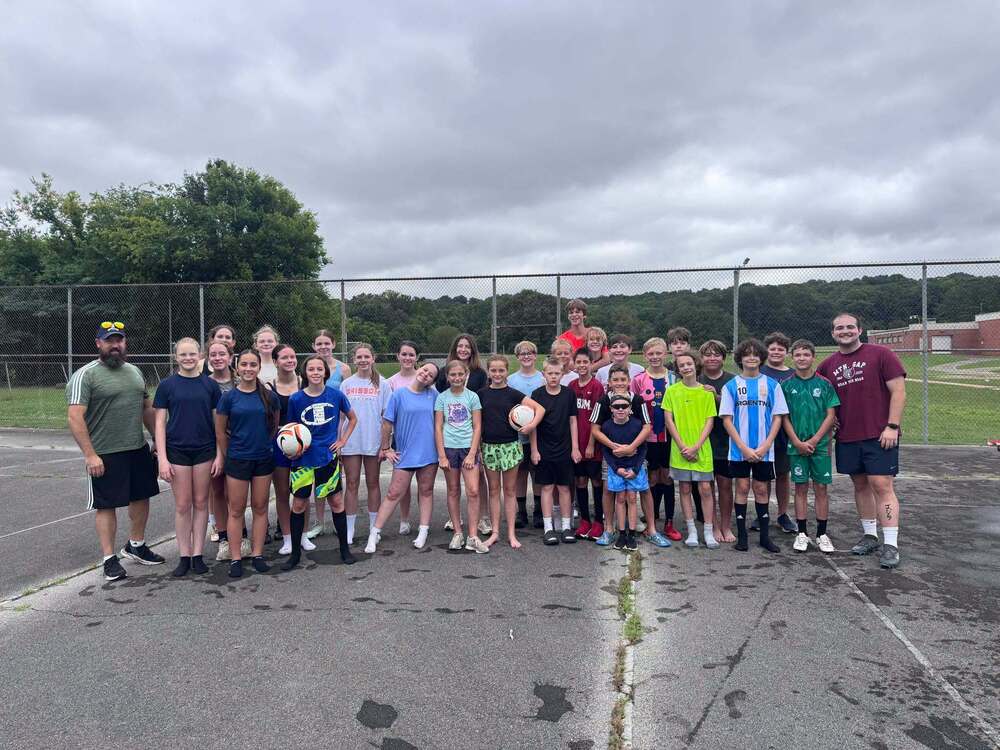 A picture of the soccer team . They are standing on asphault and there is a chain linked fence behind them. 