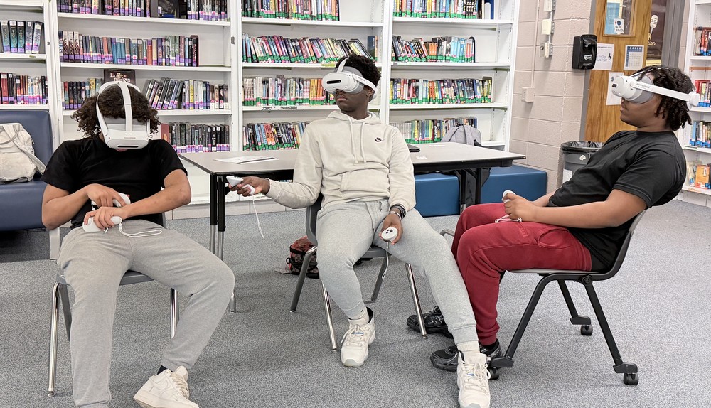 Three 8th-grade Chapman Middle School students are seated in a library, fully immersed in wearing white Prisms VR headsets and holding motion controllers. The student on the left, in a black shirt and grey pants, looks downward; the middle student, in a grey Nike hoodie and grey pants, looks slightly right; the student on the right, in a black shirt and red pants, looks far right, all actively engaged with the virtual lesson.
