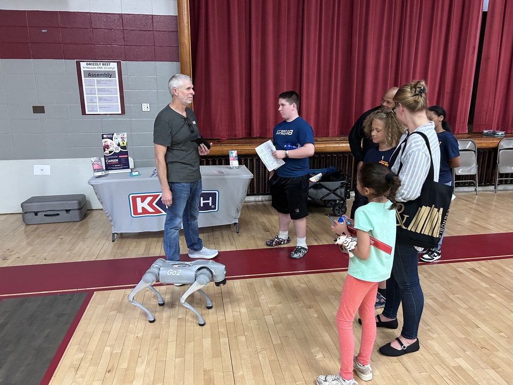 A man standing in a school gym demonstrating a small robotic dog to a group of students and adults gathered around him.
