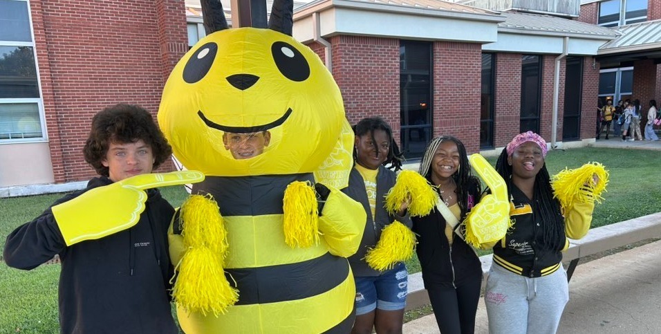 four students from yellow house greet cars in carline. One student is dressed as an inflatable bee.