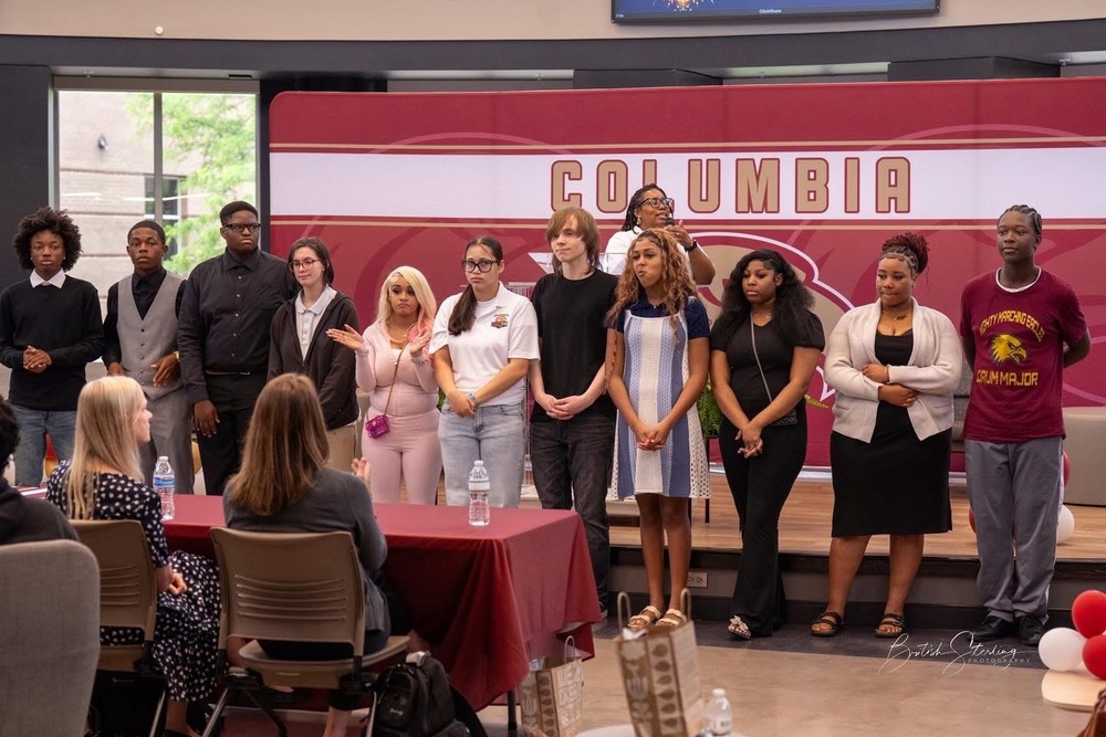 11 students standing in front of stage with Curriculum Specialist on microphone behind them and two women sitting at a table with red cloth in front of them
