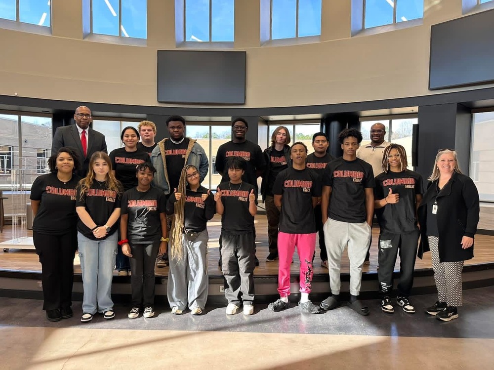 The members of the Student Leadership team at Columbia dressed in black tshirts with Columbia written in red pose for a picture with Superintendent Suuton on the left back and pricipal Richey on right in back with lady from central offic onnfar right in black sweater