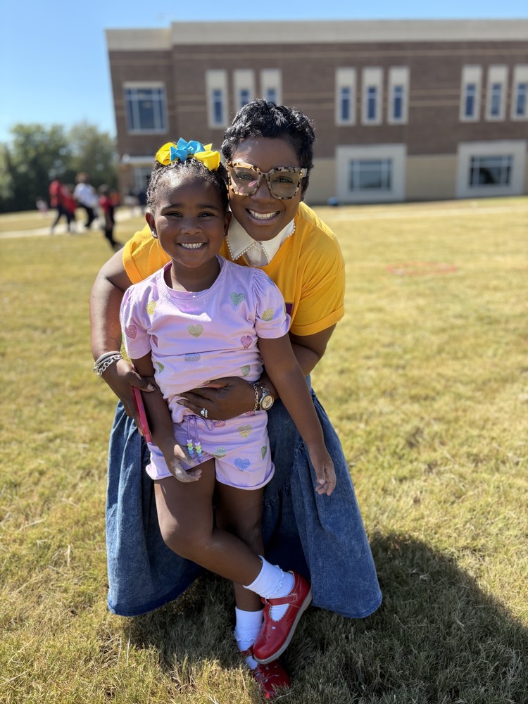 A smiling young student poses with a smiling staff member on the school lawn during an outdoor school event.
