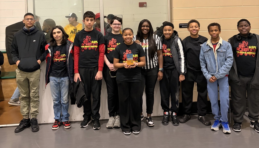 A diverse group of young individuals in a gym, joyfully posing after winning a design award and qualifying for State.