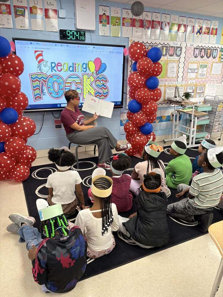 A teacher sits on the rug reading a book aloud while students gather around and listen attentively