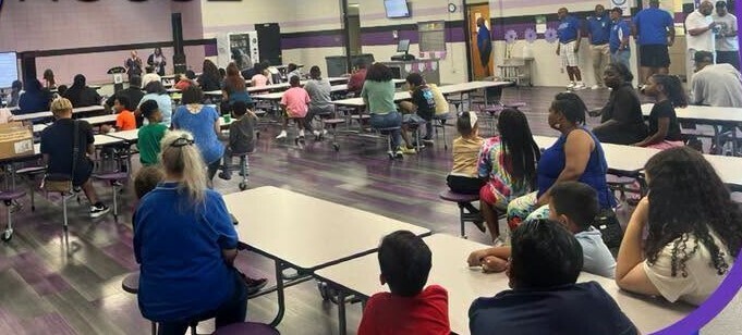 Families are seated in the cafeteria and all are facing the screen at the front of the room