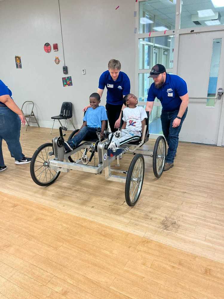 Two students sit in a UAH Moon Buggy while two UAH engineering students push it, demonstrating teamwork and STEM learning.