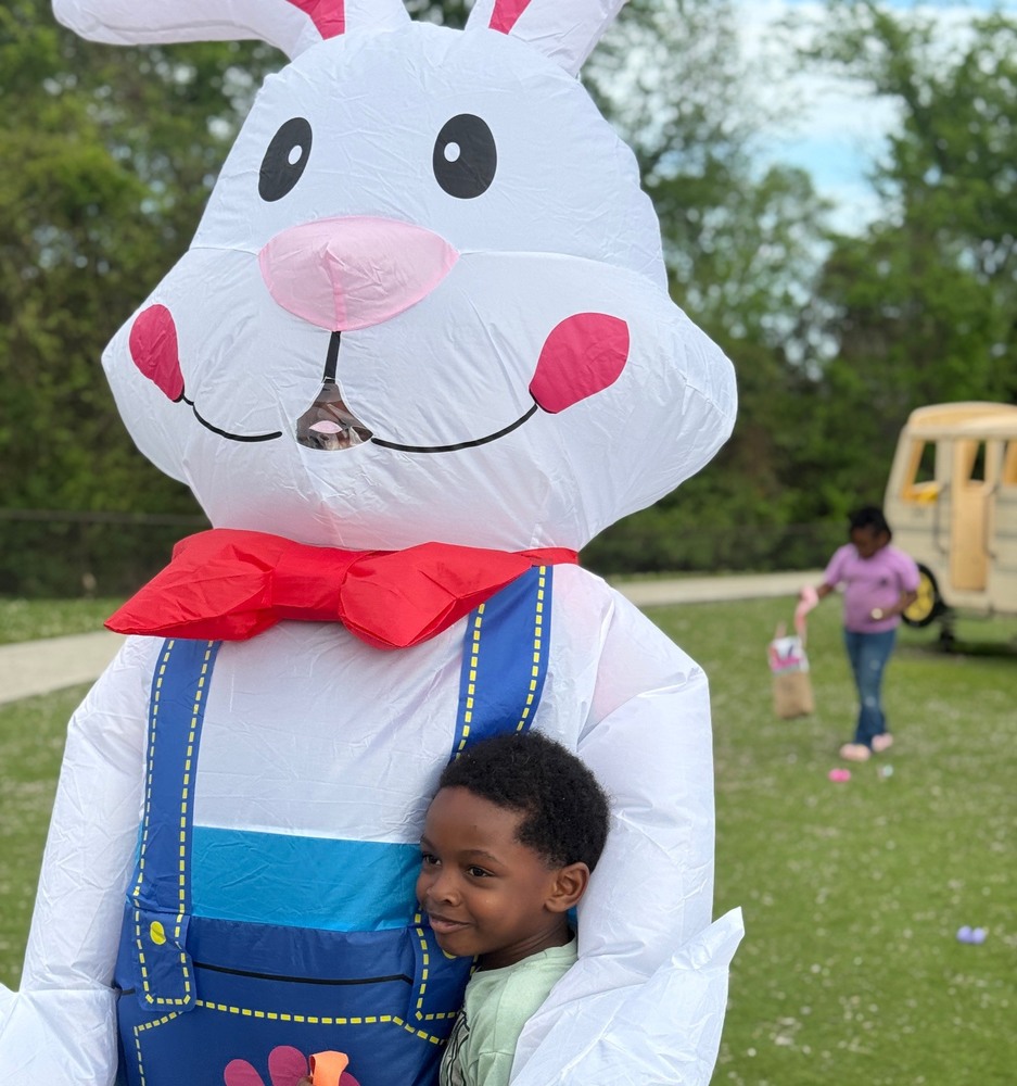 A smiling young child hugs a large inflatable Easter bunny decorated with a red bow tie and blue overalls during an outdoor egg hunt. In the background, another child walks across the grassy field holding a basket, with scattered colorful eggs and a small play structure nearby.