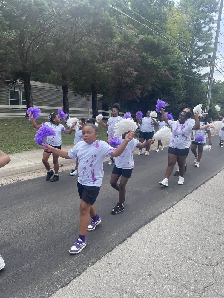Seven cheerleaders are marching through the streets, waving purple and white pom poms. They are wearing purple and white tie-dyed shirts and black shorts.