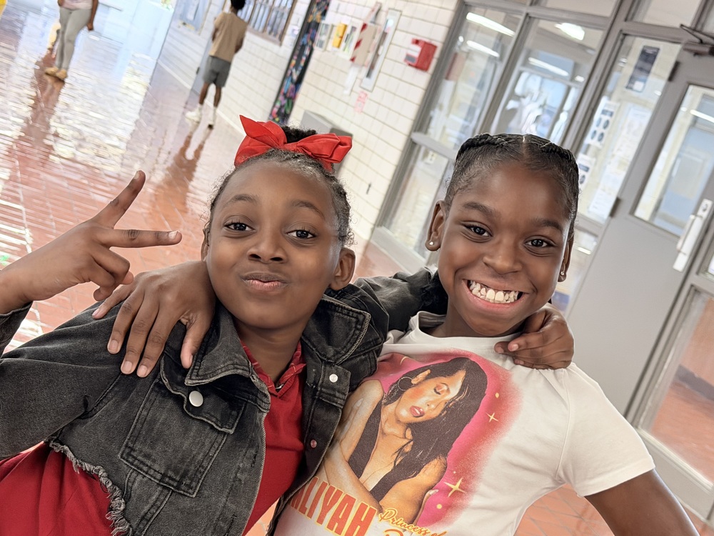 Two students are sharing their excitement with arms draped across others shoulders in the hallway as they prepare to enter the perfect attendance celebration. 