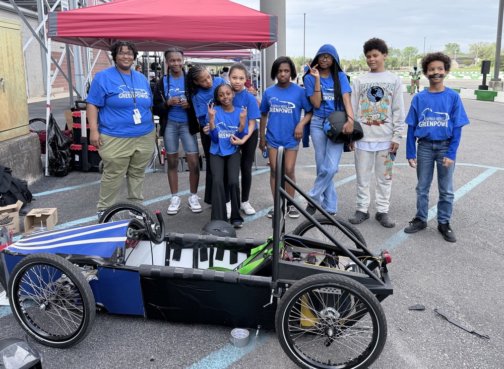 The Chapman Middle School GreenPower team and their sponsor, Ms. Jones, pose for a group photo behind their open-frame electric racing car under a grey sky.