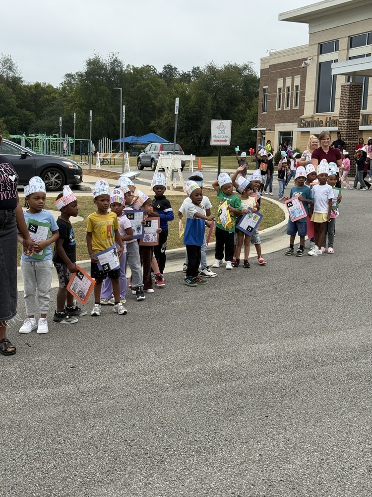 Kindergarten students participate in an alphabet parade outside Sonnie Hereford Elementary, wearing handmade alphabet hats and carrying colorful letter-themed signs and projects