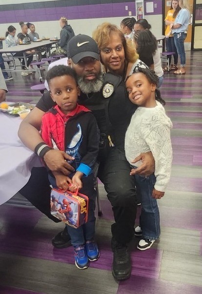A grandfather in a black ball cap and a grandmother are in a family pic with their two young grandchildren in the cafeteria.