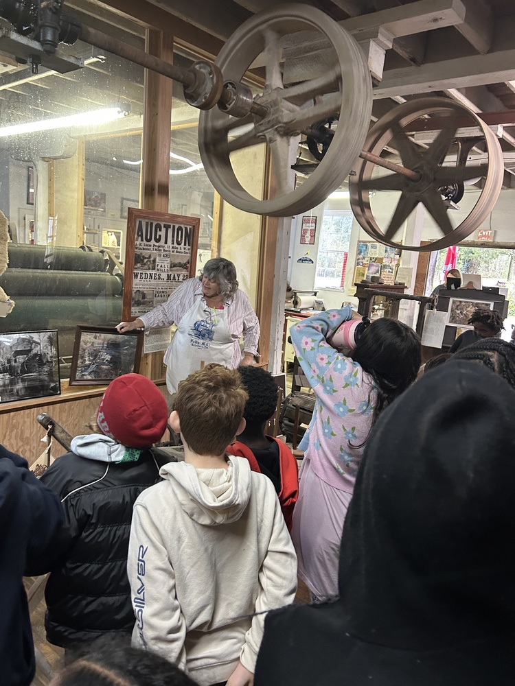 A group of students are facing a woman who is standing under large wheels  in an old mill.