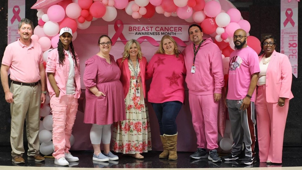 8 Staff dressed in pink and white standing in front of Pink and white balloon arch with three pink ribbons around arch with pink bulletin board paper in the middle with pink bows and breast cancer sign at the top